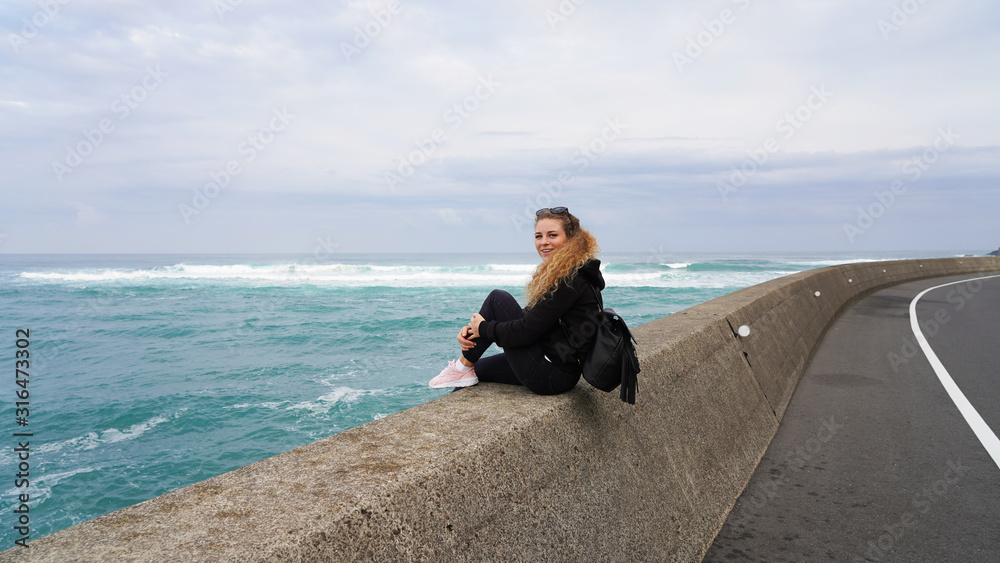 beautiful curly-haired blonde girl sitting on a breakwater near the ocean. girl looks into the distance at the pacific ocean. pacification rest in sunglasses. emotions of a young girl watching the sea