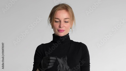 A beautiful blonde girl in black clothes puts black latex gloves on her hands. then she takes off her gloves and throws them down. Girl posing on a light background photo