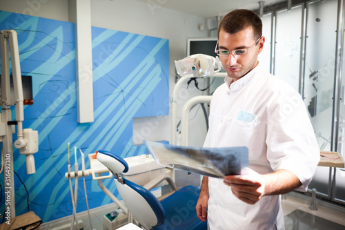 Young doctor dentist man in white uniform and glasses standing and looking at tooth picture in dental office in clinic with equipment at background. Dental health and medical care concept