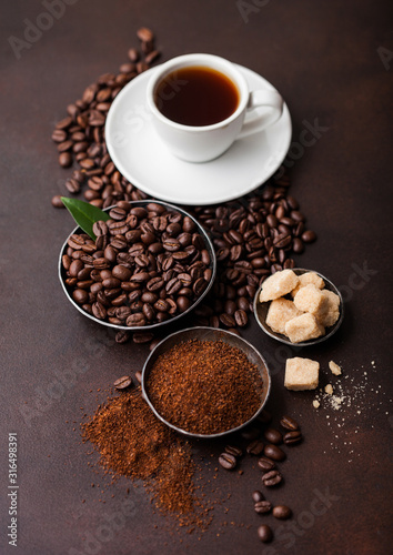 White cup of fresh raw organic coffee with beans and ground powder with cane sugar cubes with coffee tree leaf on brown background. Top view