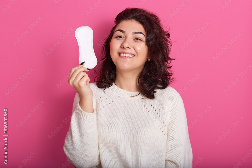 Photo of delighted charismatic young lady posing isolated over pink background in studio, raising one hand, holding hygiene pad, looking directly at camera, smiling sincerely. Menstruation concept. Photo of delighted charismatic young lady posing isolated over pink background in studio, raising one hand, holding hygiene pad, looking directly at camera, smiling sincerely. Menstruation concept.