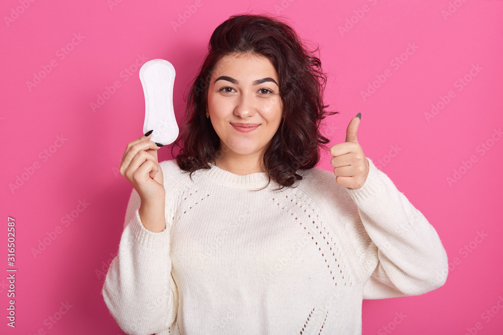 Image of smiling curly haired pretty young female making gesture, showing sign super, holding hygiene pad in one hand, making recommendation, being delighted, having menses. Menstruation concept. Image of smiling curly haired pretty young female making gesture, showing sign super, holding hygiene pad in one hand, making recommendation, being delighted, having menses. Menstruation concept.