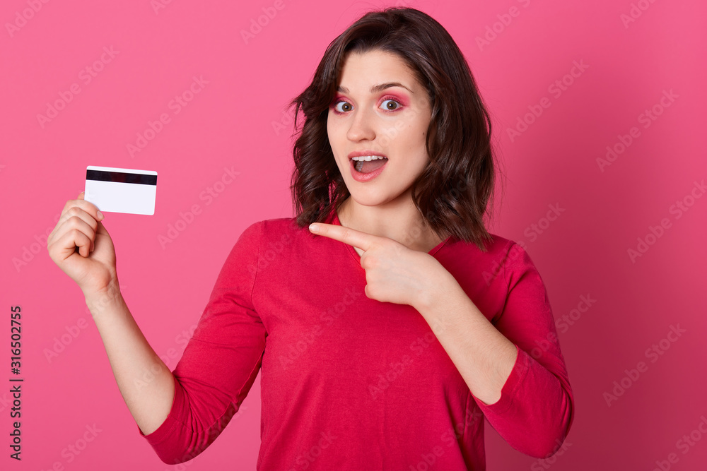Picture of emotional good looking pretty female pointing with forefinger at credit card, having surprised facial expression, opening mouth, doing shopping online. Women and purchases concept. Picture of emotional good looking pretty female pointing with forefinger at credit card, having surprised facial expression, opening mouth, doing shopping online. Women and purchases concept.