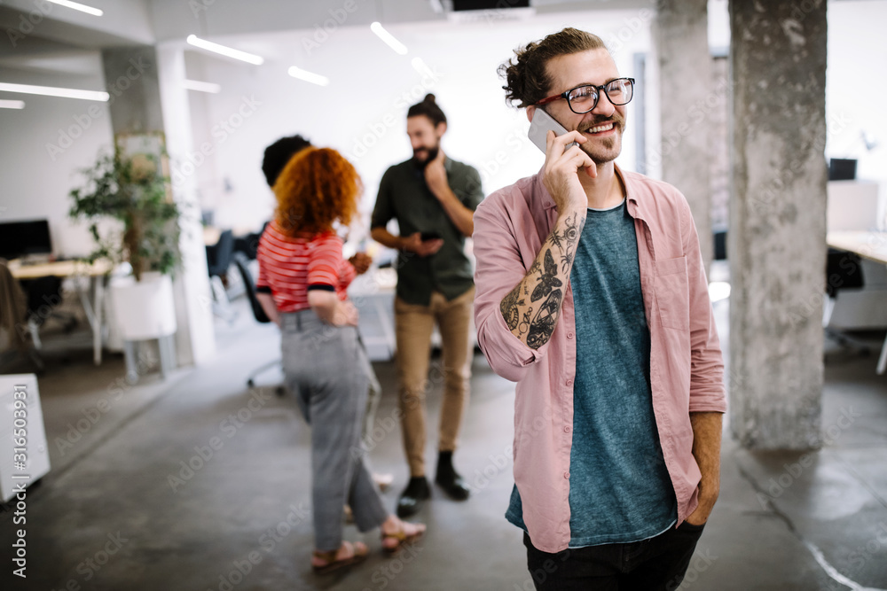 Happy young man looking at his mobile phone and smiling while his colleagues working in office Happy young man looking at his mobile phone and smiling while his colleagues working in office