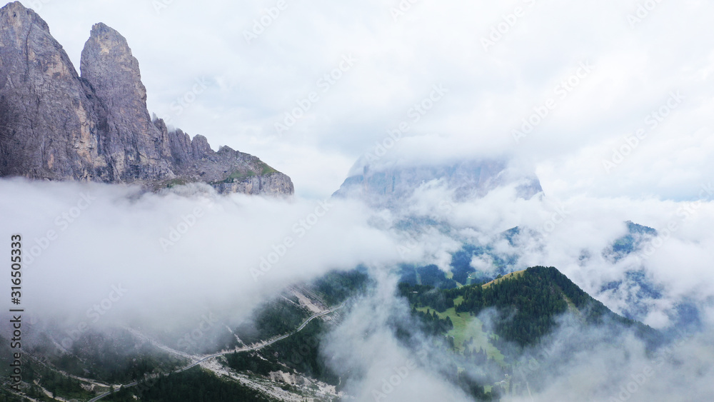 Aerial view of Dolomites in fog, low clouds. Road goes along dolomite cliffs and fir tree forest. South Tyrol, Italy. Beautiful landscape.