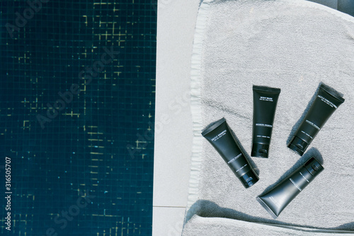 (Blank Label) Mini bottles with cosmetic products and towels beside swimming pool against sun light. Hotel amenities such as hair shampoo, hair conditioner, body wash and body lotion.