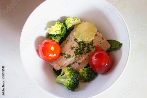 Poached salmon salad in a bowl with brocolli, tomatoes and lemon.