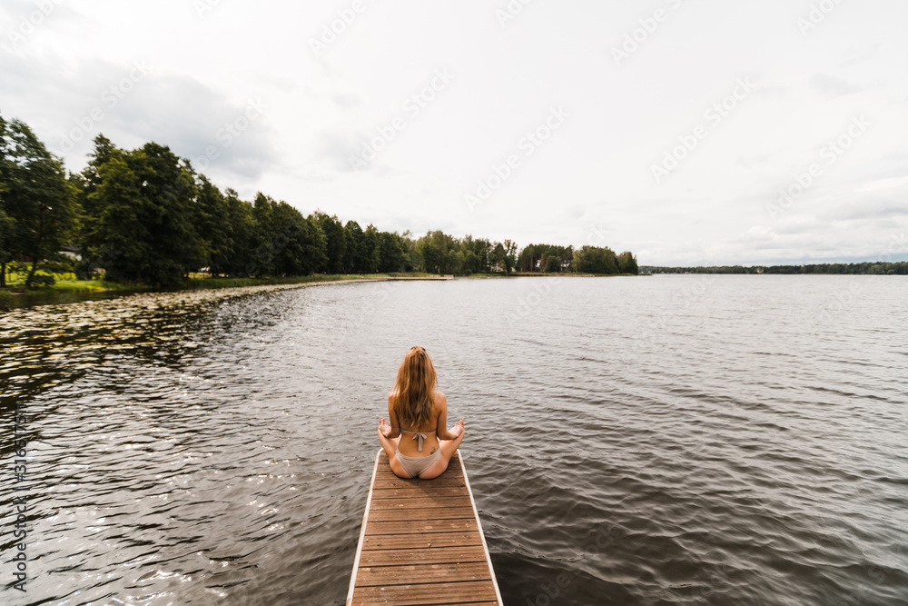 Fit slim woman practicing yoga exercises wearing mini swimsuit bikini at lake with clouds - Yoga meditation and wellness lifestyle concept - Full sitting shot