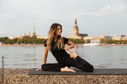 Yoga meditation and wellness lifestyle concept. Fit slim woman practicing yoga exercises near river Daugava and Vecriga old town background at sunset - Full sitting shot and meditating on rock beach