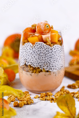 Healthy smoothie layered in glass with chia seeds in vegan coconut milk and joghurt, mixed with cereals and topped with orange persimmon and physalis fruits 