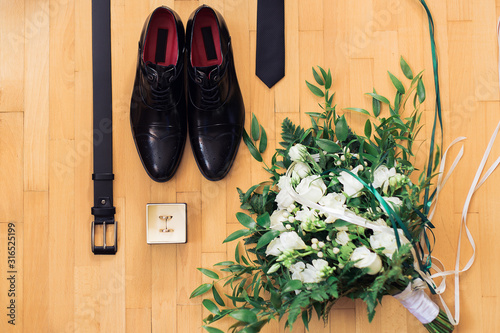 Closeup of elegant stylish black male accessorieson wooden background. Top view of belt, shoes, floral corsage, golden rings. Preparation for wedding concept. photo