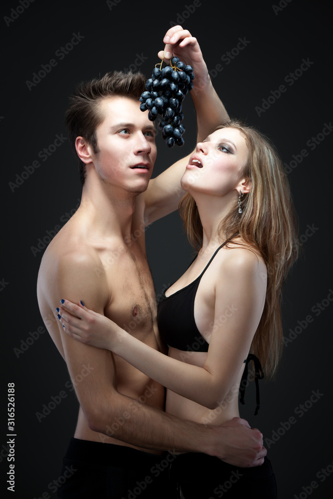 Fototapeta premium Young beautiful loving sexy couple standing and reaching for bunch of black grapes over dark grey background