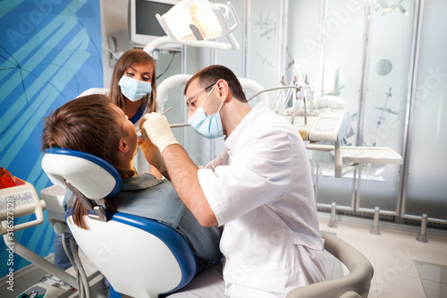Young dentist doctor man in white uniform and special mask and young nurse woman examining man patient in dental office in clinic. Dental health and medical care concept