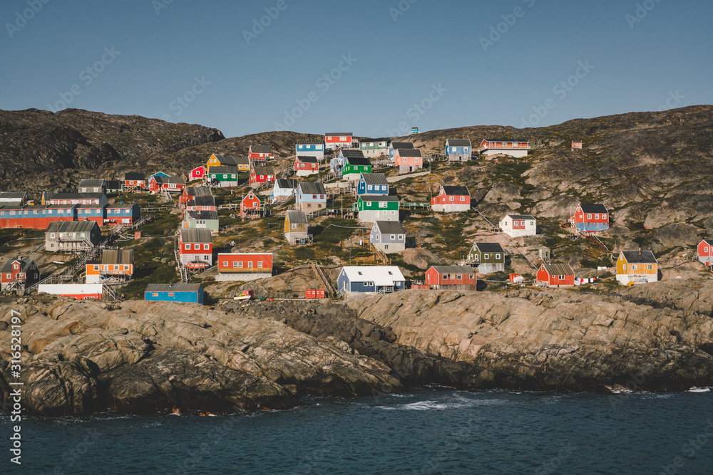 Colorful houses dot the hillsides of the fishing town of Kangaamiut, West Greenland. Icebergs from Kangia glacier in Greenland swimming with blue sky and clouds. Symbol of global warming.