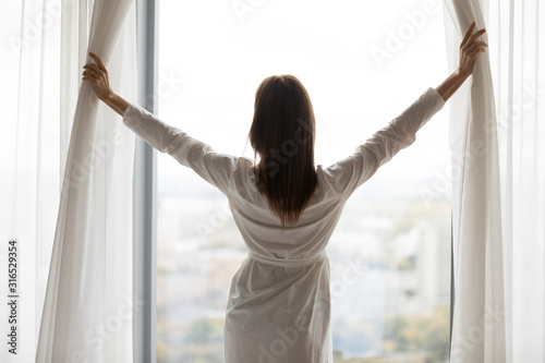 Free Download Rear view beautiful woman wearing bathrobe opening curtains in morning - GFXLoad