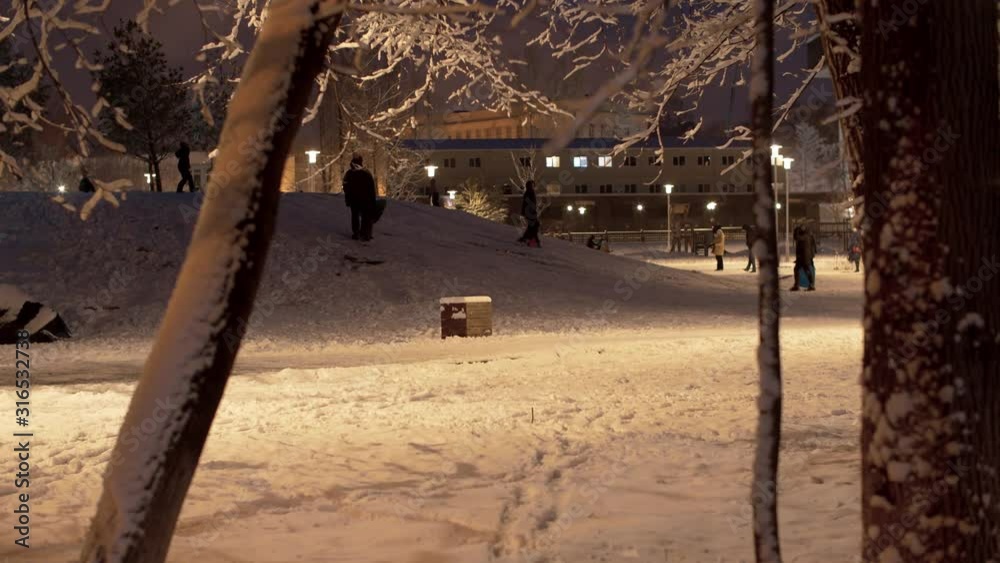 Children riding down a hill in snow covered city park. Fresh snow, magical winter evening. Unrecognizable people on the stroll after snowfall. Panorama, steadicam shot Children riding down a hill in snow covered city park. Fresh snow, magical winter evening. Unrecognizable people on the stroll after snowfall. Panorama, steadicam shot