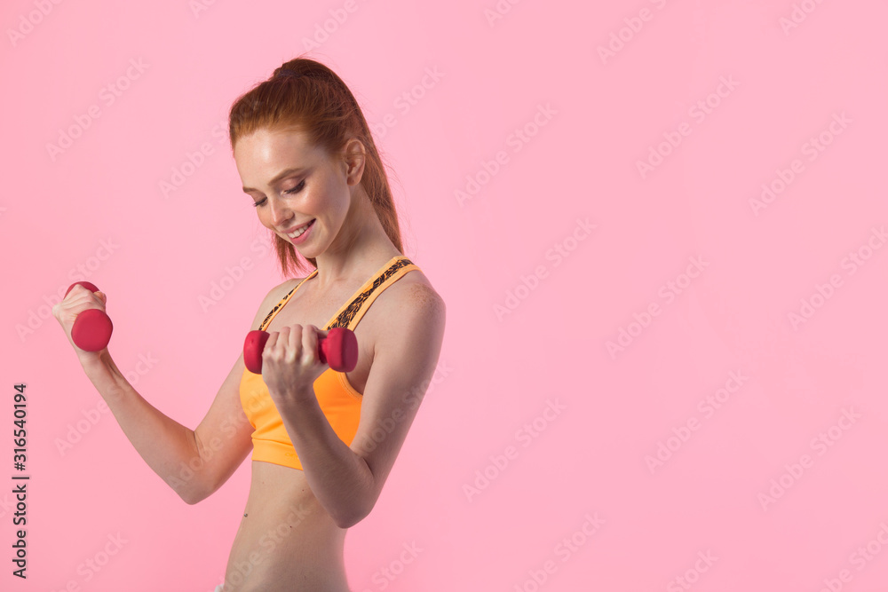 beautiful young woman with red hair on a pink background with dumbbells in hands