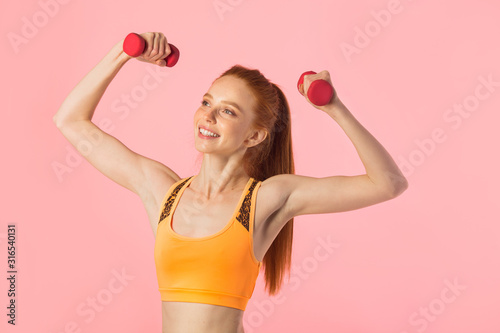 beautiful young woman with red hair on a pink background with dumbbells in hands