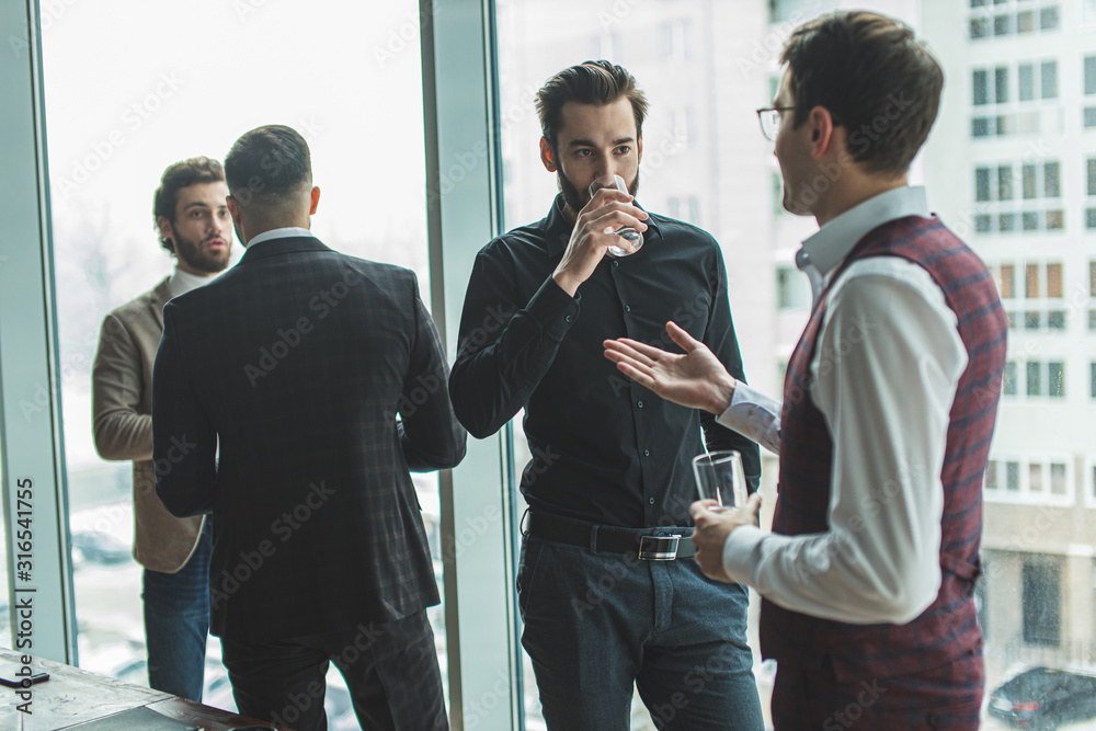 free time of caucasian colleagues at work in office, handsome men in formal wear stand next to panoramic window and have conversation. business people concept free time of caucasian colleagues at work in office, handsome men in formal wear stand next to panoramic window and have conversation. business people concept