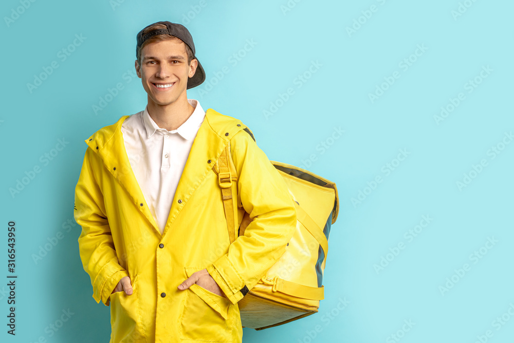 portrait of nice deliverman in yellow uniform isolated over blue background, wearing cap and carrying backpack with orders, look at camera and smile portrait of nice deliverman in yellow uniform isolated over blue background, wearing cap and carrying backpack with orders, look at camera and smile