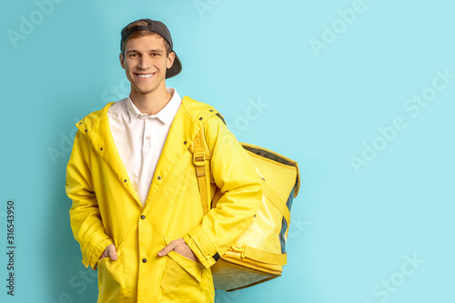 portrait of nice deliverman in yellow uniform isolated over blue background, wearing cap and carrying backpack with orders, look at camera and smile photo