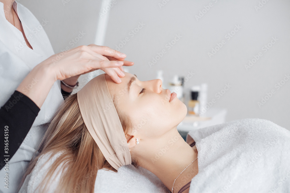 Fototapeta premium Beautician massage therapist making facial massage to a beautiful female client sitting on a couch in a cosmetology office. Skin care concept