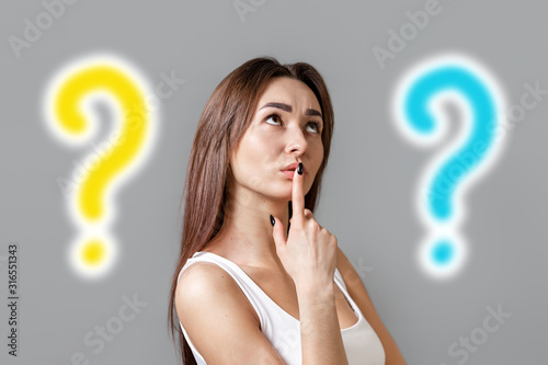 The concept of searching for ideas. Portrait of a pensive young Caucasian woman looking at the top and putting her finger to her mouth. Gray background with two question marks. Copy space