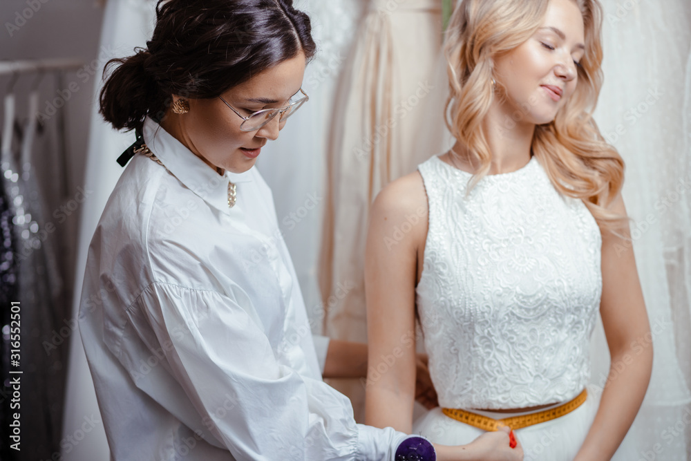 beautiful caucasian woman client and tailor in wedding boutique, taking measures from body for creation new dress, preparing for a wedding beautiful caucasian woman client and tailor in wedding boutique, taking measures from body for creation new dress, preparing for a wedding