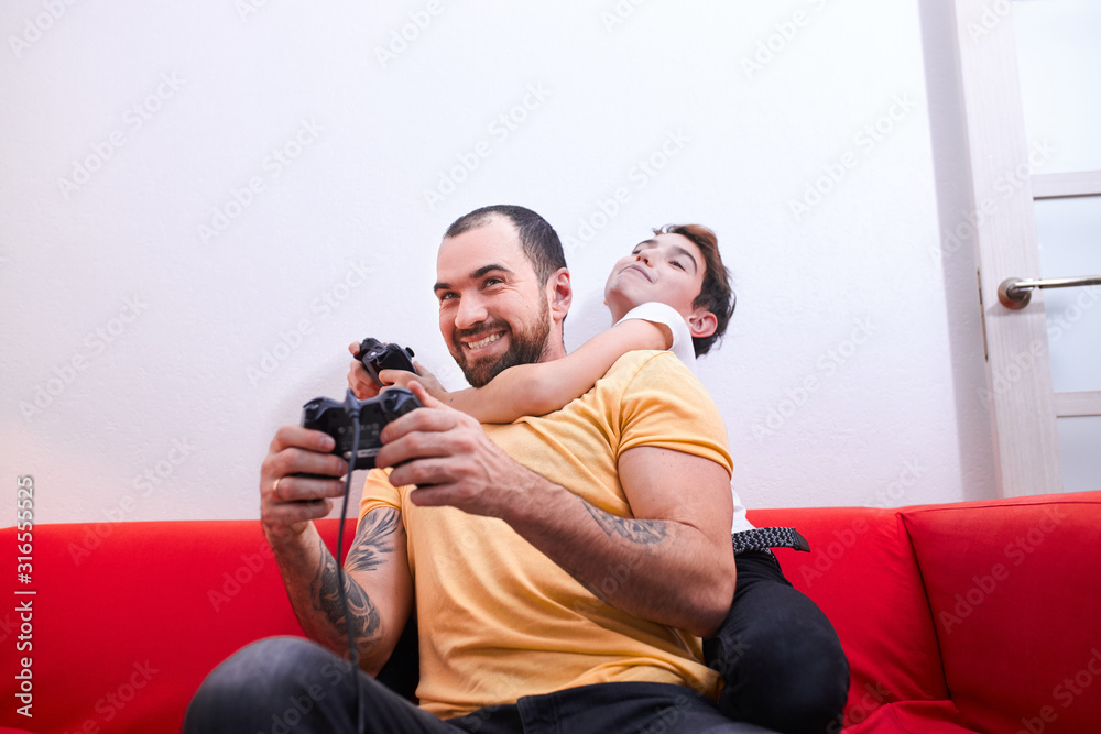 Φωτογραφία young caucasian man with beard sit with son on sofa and play video game together, holding joysticks, wearing casual t-shirt