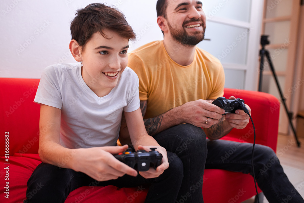 joyful playful father and son playing video game sitting on red sofa, happy parent and kid boy enjoy spending time together - obrazy, fototapety, plakaty 