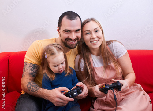 young parents with kid girl sit playing video game together, holding joysticks, wearing casual clothes. modern technologies, happy family concept