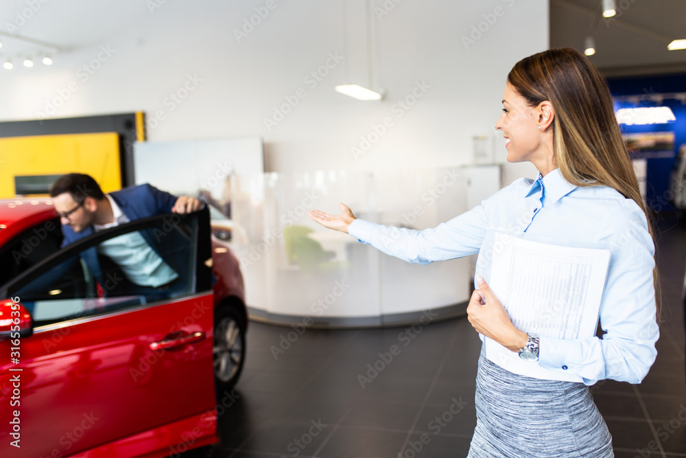 Saleswoman at the dealership showroom talking with customer and helping him to choose a new car for himself. Saleswoman at the dealership showroom talking with customer and helping him to choose a new car for himself.