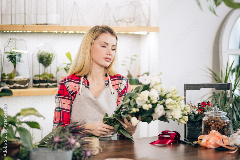 flowers composing and decoration by young lady with blonde hair, wearing red checkered shirt, isolated in light room full of green plants. botany, flowers concept