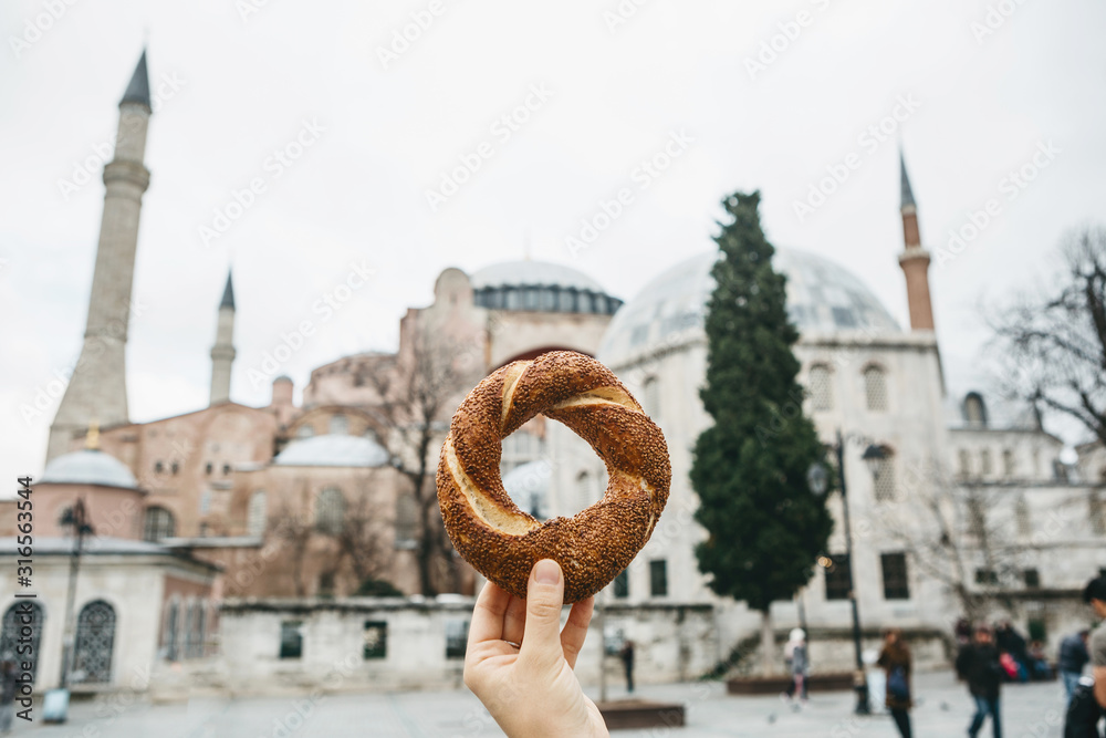 Fototapeta premium A person holds a traditional Turkish pretzel Simit against the background of the landmarks of Hagia Sofia in Istanbul.