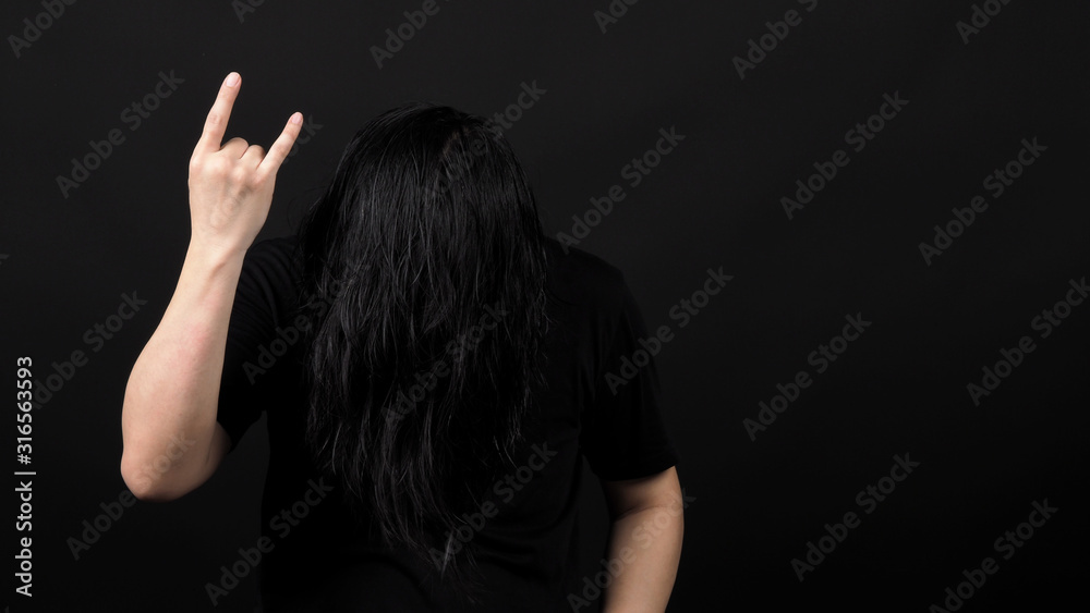 Obraz premium Rocker man long hair and making hand sign of rock and roll and shaking head with black color t-shirt and dark background paper in studio which represent style of music such as punk or heavy metal
