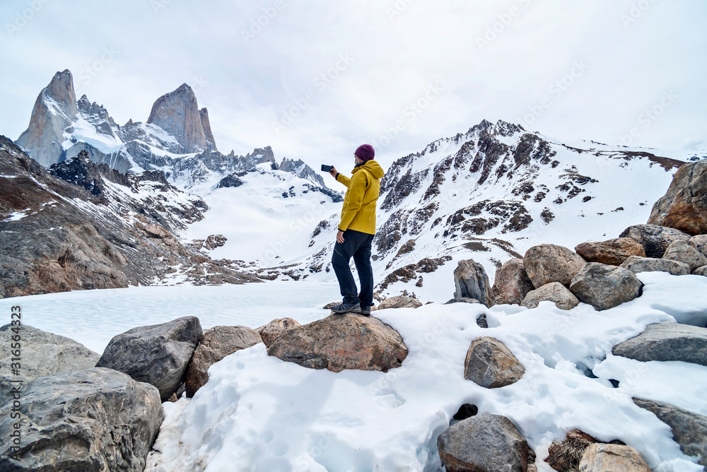 A hiker with a yellow jacket taking a photo on the base of Fitz Roy Mountain in Patagonia, Argentina