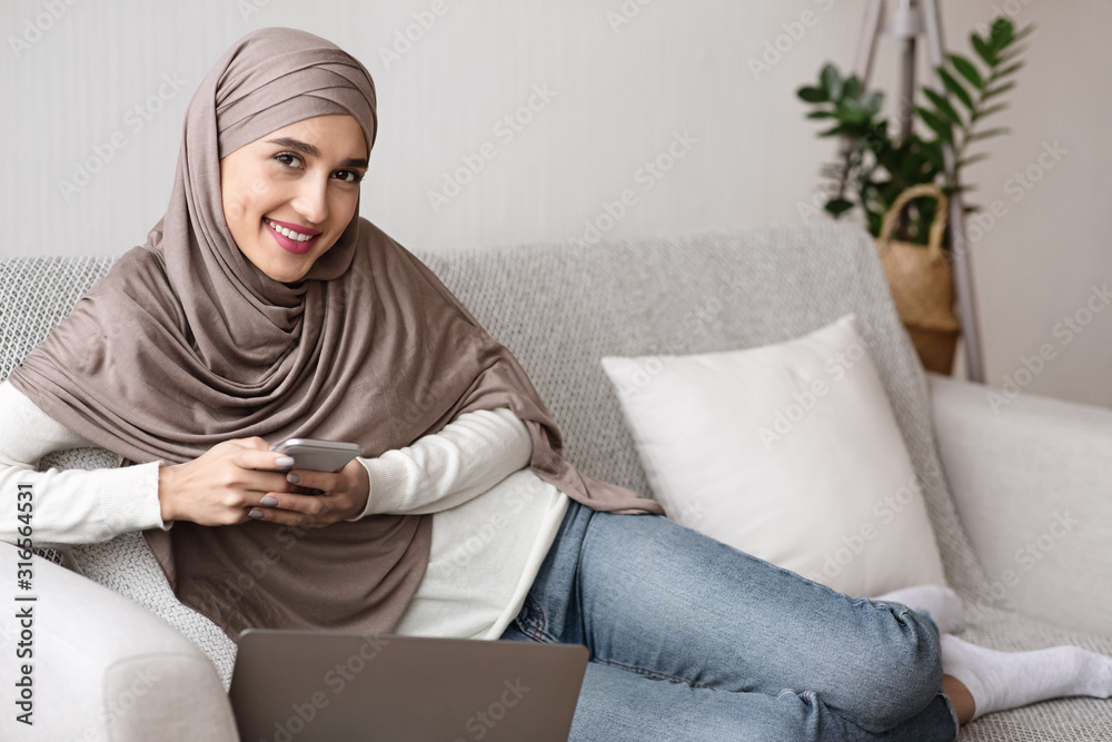 Cheerful muslim girl in hijab spending time with smartphone at home