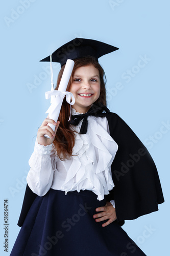 Vertical view. Frontal portrait of a happy little girl in graduation cap standing and showing a success moment, over blue background. photo