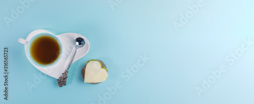Cup of tea and silver spoon on white saucer in heart shape and sandwich in heart shape on light blue background. Banner.