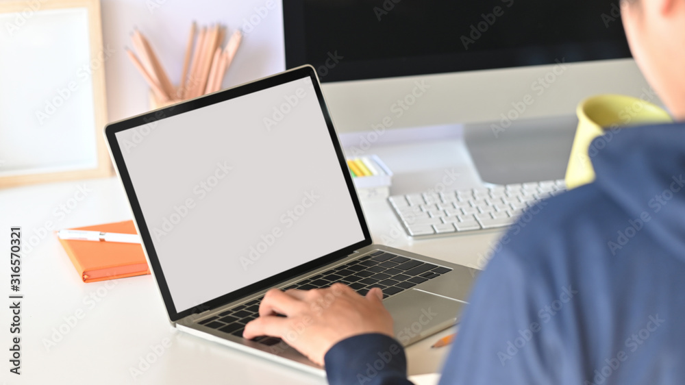 Behind shot of smart programmer holding coffee cup and typing on white blank screen laptop while sitting over his black screen monitor on modern working desk. Behind shot of smart programmer holding coffee cup and typing on white blank screen laptop while sitting over his black screen monitor on modern working desk.