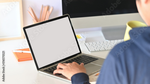 Behind shot of smart programmer holding coffee cup and typing on white blank screen laptop while sitting over his black screen monitor on modern working desk.