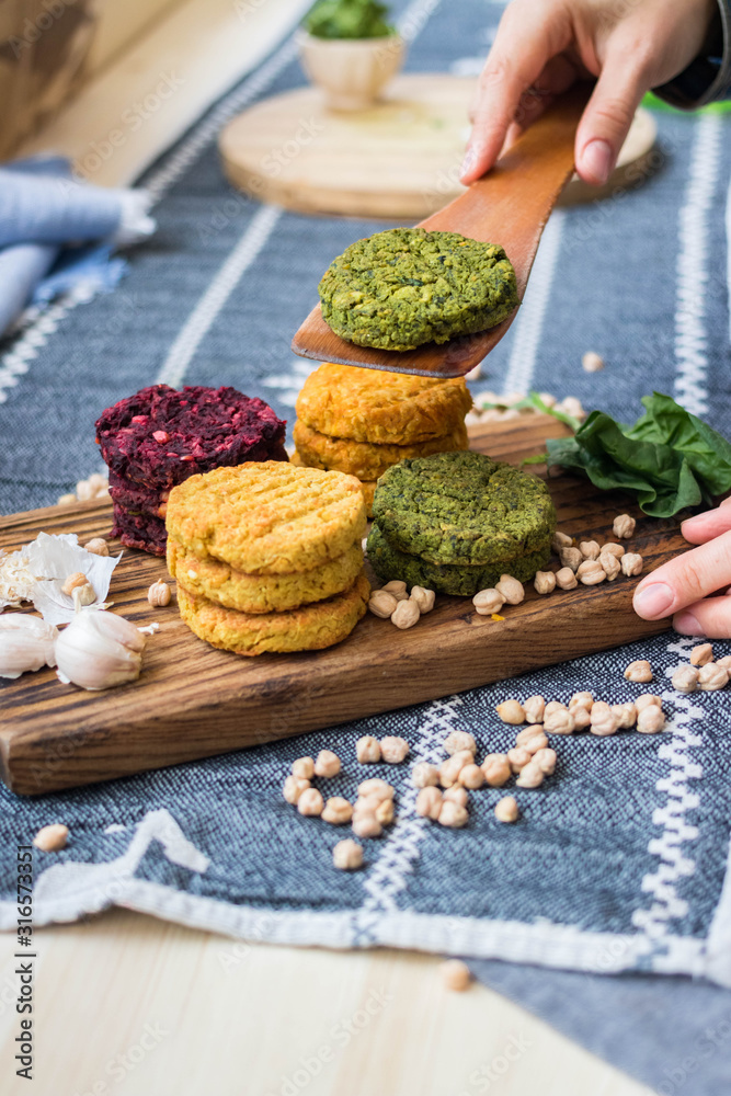 Vegan chickpea burgers cutlets or patties. Healthy vegan diet food. Woman hands holds mixed vegetables yellow pumpkin, orange carrot, green spinach and red beet bean cutlets with wooden spatula