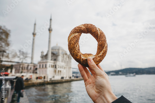 A man holds in his hand a traditional Turkish bagel called Simit against the background of the Ortakey Mosque in Istanbul in Turkey
