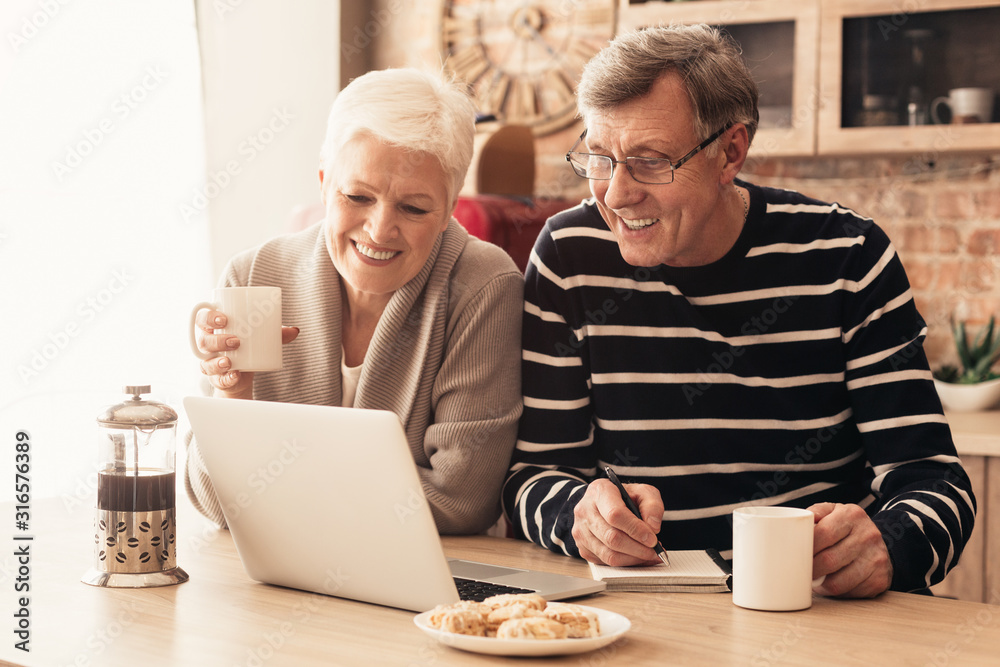 Cheerful senior couple watching news on laptop online at kitchen - obrazy, fototapety, plakaty 