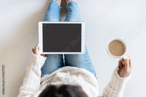 Smiling pretty woman in white sweater using tablet computer while holding a cup of coffee sitting in the living room at home. Top view, copy space.