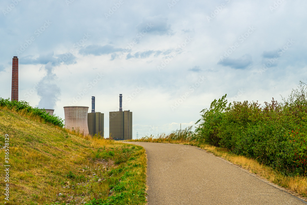 View across meadows, pathway and bushes to a power plant with steaming cooling tower on the horizon View across meadows, pathway and bushes to a power plant with steaming cooling tower on the horizon