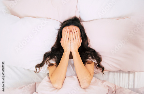 Insomnia! It's so bad. Close-up photo of a young woman with curly brown hair, who is laying in her bed on pink pillows, covering her face with palms.