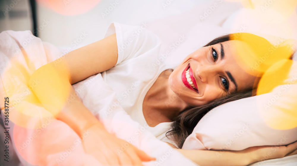 Fototapeta premium Taking a catnap. Close-up photo of a good-looking woman, who is laying in her bed, hugging a pillow and smiling, while looking in the camera.