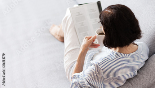 Middle-aged brunette woman with glasses on the gray sofa reading magazine with cup of coffee, soft focus comfort concept of loneliness.