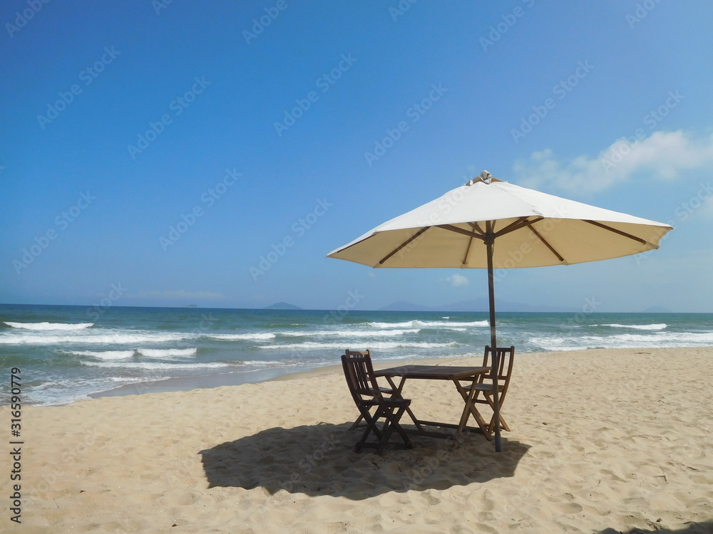 A secluded sandy beach scene with white breakers in background, foreground of wooden table and chairs with large cream parasol.